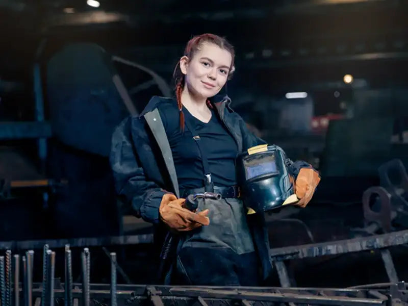 Eine Schweißerin in Ausrüstung mit ihrem Schweißhelm in der Hand, die in einer Werkstatt steht. Sie hat den perfekten Schweißhelm für sich gefunden.